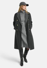 Grijze wolmix jas met houndstooth patroon, dubbele knopen, gouden knopen, gecombineerd met een grijs coljurk en zwarte laarzen tot de knie.