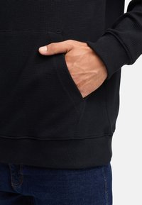 Sort ribbet stof hoodie med en kængurulomme. Blød tekstur med en kantet bund og manchetter. Båret med mørkeblå jeans.