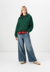 Jeune femme se tenant debout, portant un sweat à capuche vert, une chemise à carreaux rouge et noir, un jean large bleu et des baskets beige, contre un fond blanc uni.