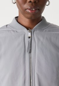 Světle šedá bomber bunda s žebrovaným límcem, předním zipem a hladkým povrchem. Obsahuje detail s kovovým zipem pro bezpečné uzavření.