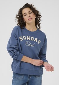 Mlada žena s kovrčavom kosom koja nosi plavu duksericu s natpisom "SUNDAY Club", podešava rukav, stojeći ispred jednostavne svijetle pozadine.