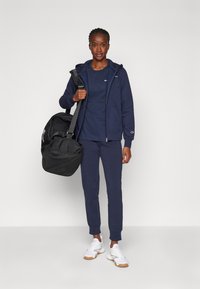 Marine sportoutfit met een rits trui, T-shirt met ronde hals en tapering sweatpants. Zwarte sporttas en witte sneakers met rubberen zolen.
