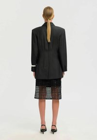 Ženska z blond čopom, oblečena v prevelik črn blazer, črno mrežno krilo in črne pete, ki stoji ob preprosti beli ozadju.