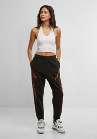 Svarte joggebukser med røde sidestreker, paret med en hvit kort topp. Modellen har på seg hvite sneakers med grå detaljer.