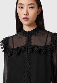 Schwarze Bluse mit transparenten Materialien, Rüschendetails, Spitzenbesatz, hohem Kragen und einem Reißverschluss vorne. Ein subtiler Glanz sorgt für Textur.