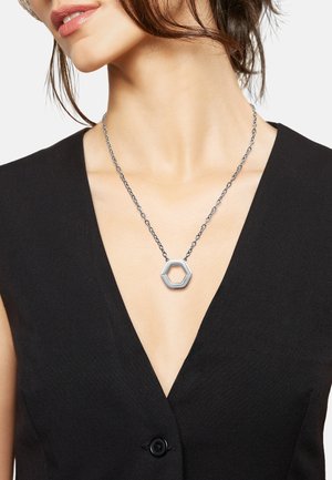 Pendente esagonale argento su una collana a catena testurizzata, indossato su un top nero a scollo a V profondo, che mette in mostra il design geometrico del pendente.