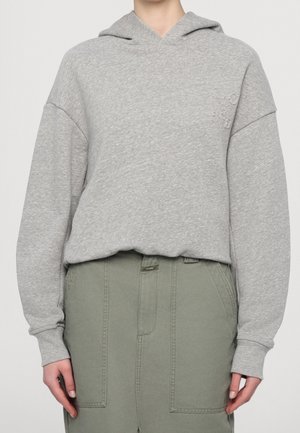Personne portant un sweat à capuche gris clair avec un logo discret en relief et un pantalon taille haute vert olive avec de grandes poches avant.