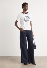 Maglietta bianca a maniche corte con colletto a coste blu marino e stampa grafica, abbinata a jeans a gamba larga in denim blu scuro e una piccola borsa nera.