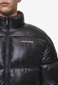 Sort, polstret pufferjakke med skinnende, quiltet tekstur. Har høj krave, frontlynlås og det prydede "PEGADOR" logo på brystet.