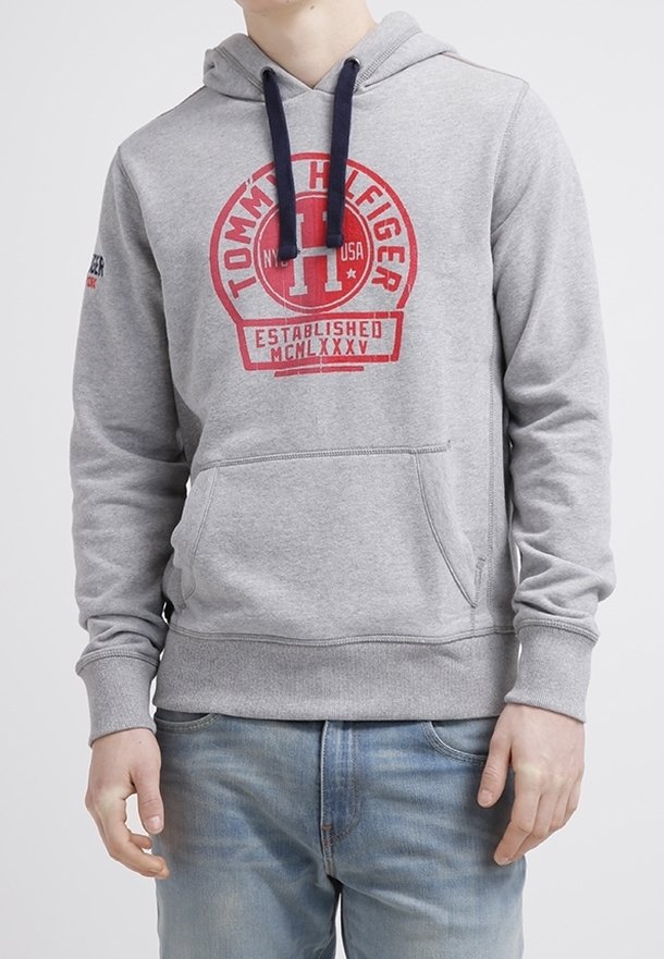Personne portant un sweat à capuche gris Tommy Hilfiger avec un logo rouge et des cordons bleus, associé à un jean bleu clair, debout devant un fond uni.