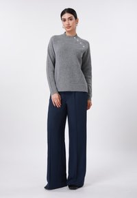 Pull gris côtelé avec un col montant et quatre boutons décoratifs sur l'épaule, associé à un pantalon large bleu marine.