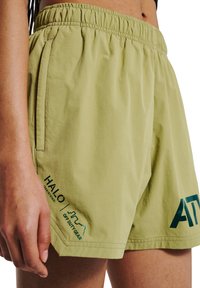 Hellgrüne Shorts mit elastischem Bund, die eine glatte Textur aufweisen. Am linken Bein sind grüne gestickte Logos für 'HALO' und 'ATV' zu sehen.