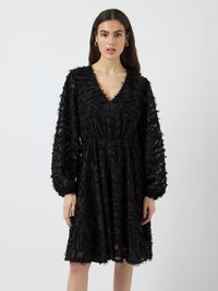 Woman wearing a knee-length black dress with textured pattern, long puffed sleeves, and a V-neckline, standing against a plain background.