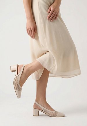 Kobieta w beżowych siatkowych sandałach na obcasie typu blok oraz lekkiej, przezroczystej, jasnobeżowej sukience midi, pozująca na jednej nodze.