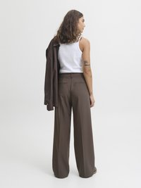 Calças largas castanhas com uma textura suave, combinadas com uma t-shirt branca. Uma jaqueta castanha a condizer está pendurada sobre o ombro.