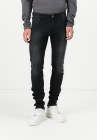 Homme portant un jean slim noir délavé, des baskets blanches et une chemise grise à manches longues, debout devant un fond blanc.