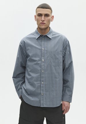 Joven con una camisa de cuadros azul y negra de manga larga abotonada, y pantalones negros, de pie frente a un fondo claro y sencillo.