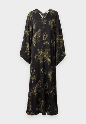 Kvinde med langt mørkt hår iført en sort maxikjole med grønne og beige botaniske print og sorte høje hælede sandaler, stående mod en hvid baggrund.