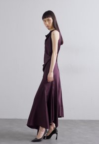 Lunga vestito senza maniche in satin viola scuro con un design drappeggiato. Corpino aderente che si trasforma in una gonna fluente. Indossato con tacchi alti neri.