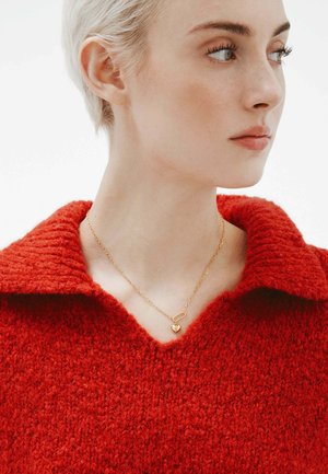 Collar de oro con un charm en forma de corazón, llevado sobre un suéter de lana gruesa de un rojo brillante con un cuello ancho y una superficie texturizada.