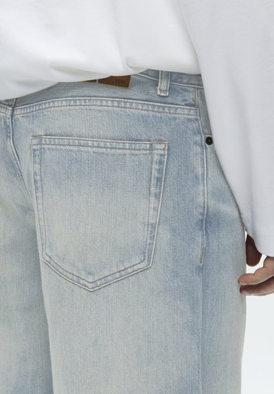 Svetlomodré rifľové džínsy s viditeľným šitím na vrecku, nosené s bielou dlhorukávovou košeľou a voľne položenou rukou pri boku.