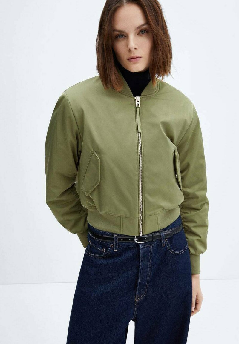 Frau mit braunen Haaren trägt eine olivgrüne Bomberjacke über einem schwarzen Top und dunkelblauen Jeans mit einem schwarzen Gürtel, steht vor einem schlichten hellen Hintergrund.