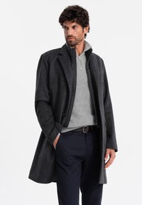 Abrigo largo gris oscuro con un patrón texturizado, usado sobre un suéter de cuello con cremallera gris claro. Combinado con pantalones azul marino, que cuentan con un detalle de botón.