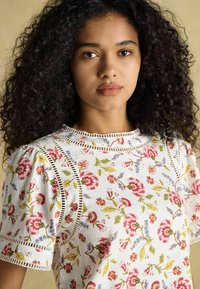 Biała bawełniana koszula z kwiatowym wzorem w kolorach różowym, żółtym i niebieskim. Posiada bufiaste rękawy oraz koronkowe wykończenie przy szwach.