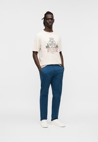 Μπεζ βαμβακερό μπλουζάκι με γραφικό τύπωμα και κοντά μανίκια, σε συνδυασμό με μπλε παντελόνια. Το μοντέλο φοράει λευκά αθλητικά παπούτσια, με τα χέρια στις τσέπες.