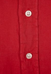 Rood linnen overhemd in close-up met een knoopsluiting en twee witte viergaatjesknopen, textiel met een zichtbare weefpatroon.