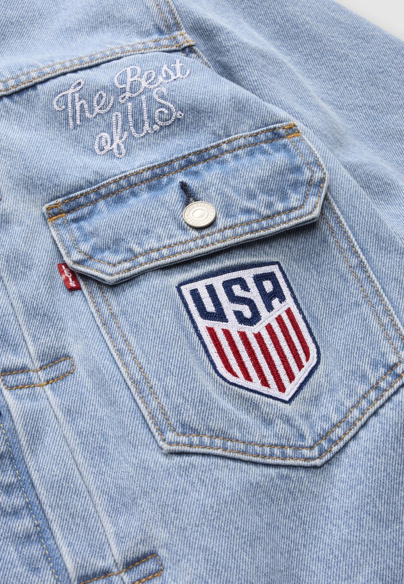 Poche de veste en denim bleu clair avec bouton, étiquette rouge « Levi's », texte brodé « The Best of U.S. » et écusson bouclier des États-Unis avec des rayures rouges.