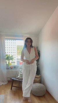 Salopetă albă din in cu decolteu în V, butoni frontali și picioare largi. Textură moale, croială lejeră, prezentată într-un spațiu interior bine iluminat cu plante.