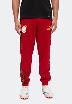 Homme portant un pantalon de sport rouge avec le logo de l'équipe sur la cuisse gauche, "KING" et le logo Puma sur la cuisse droite, des bandes latérales à motifs et des crampons blancs.