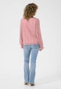 Light pink blouse with diamond lace pattern, long sleeves, and button back detail, paired with light blue flared jeans and beige heels.