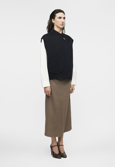 Γυναίκα που φοράει μαύρο αμάνικο γιλέκο με μεγάλη τσέπη, λευκό πουκάμισο με μακριά μανίκια, καφέ μίντι φούστα και μαύρα παπούτσια με τακούνι, στέκεται προφίλ.