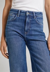 Siniset farkut, joissa on korkea vyötärö, klassinen suora lahje ja hienovarainen kuluminen. Viisi taskua, nappikiinnitys ja vyölenkit.