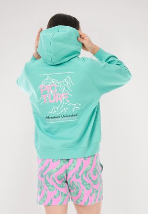 Türkisfarbener Hoodie mit einem Berggrafik und pinkem Text auf der Rückseite, kombiniert mit pinken Shorts mit grünen wirbelnden Mustern.