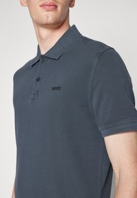 Polo shirt gris foncé avec un tissu texturé, un col à trois boutons et un petit logo noir sur la poitrine. Manches courtes avec des poignets repliés.