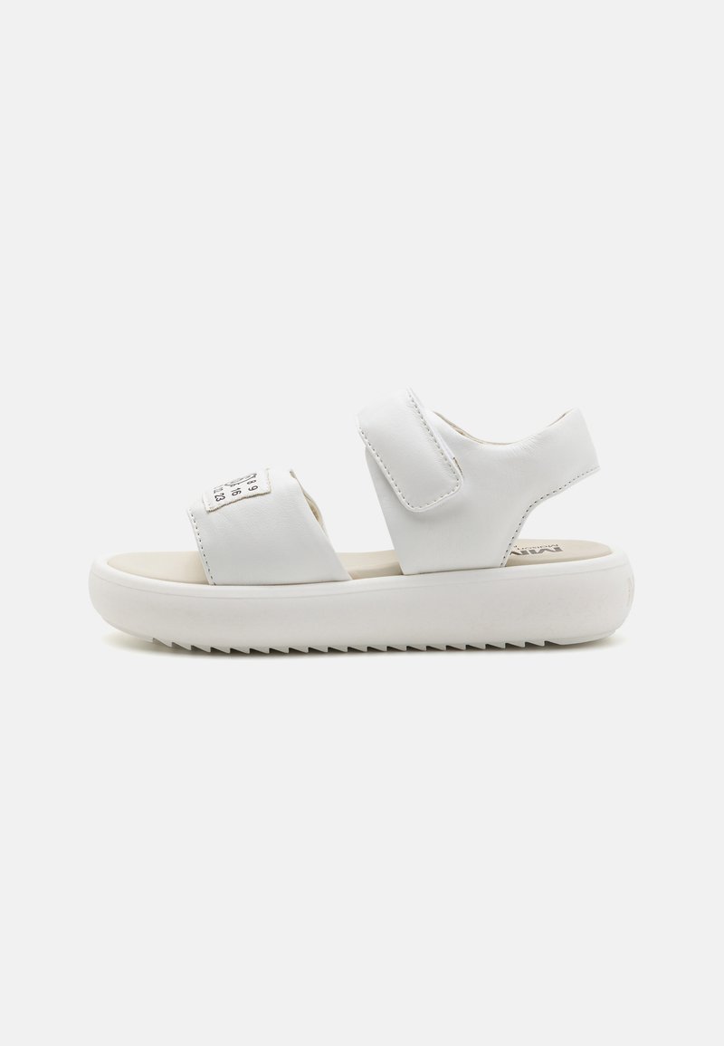 Vita sandaler gjorda av mjukt syntetiskt material med justerbara velcro-remmar, en vadderad innersula och en texturerad gummisula.