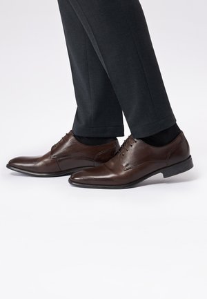 Brown leather dress shoes with a sleek design, pointed toe, and lace-up closure. Worn with dark trousers and black socks.