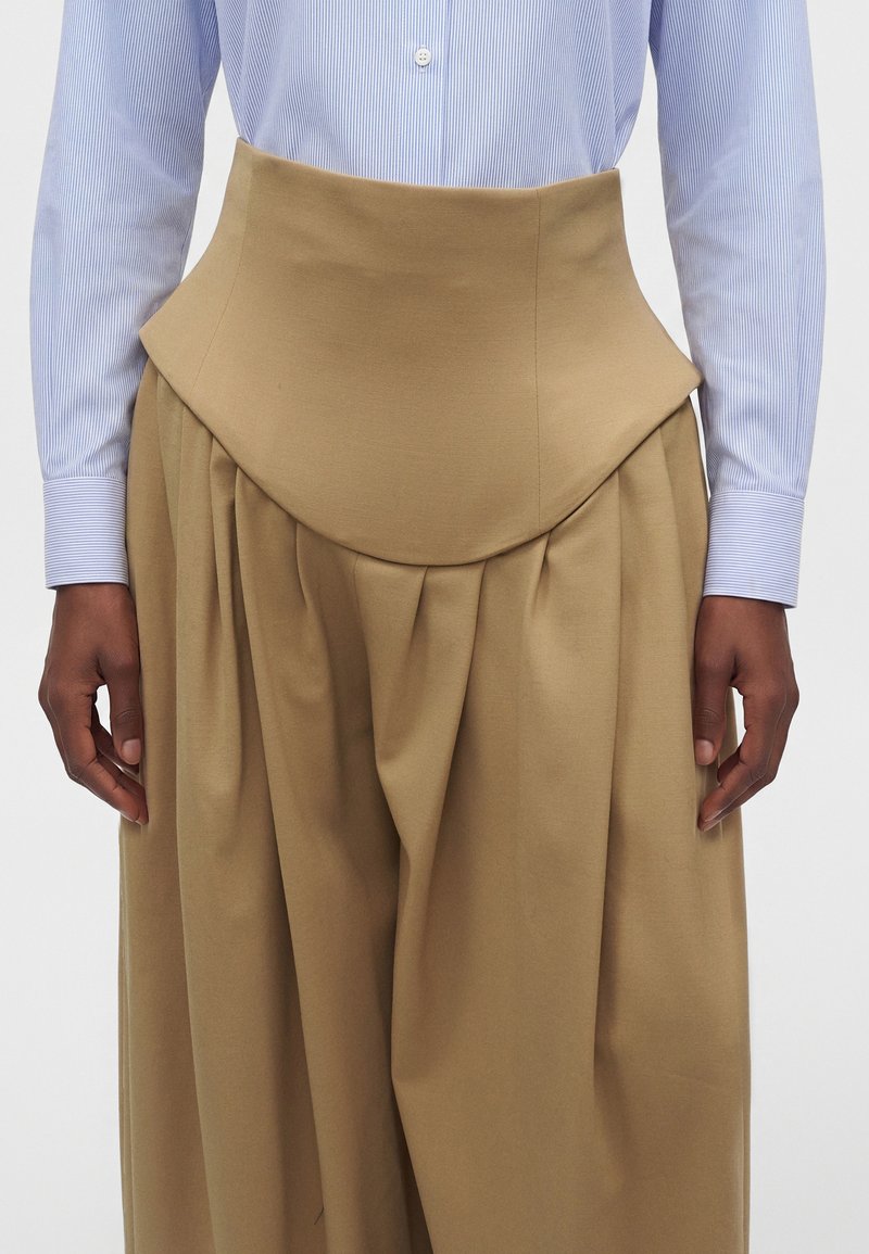 Personne portant un pantalon camel plissé taille haute, associé à une chemise bleu clair à manches longues boutonnée. Les mains reposent sur les côtés.