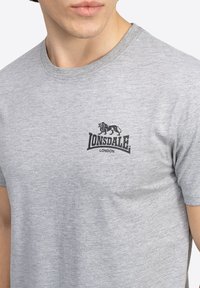 Pelēka kokvilnas T-krekls ar melnu grafisko logo, kurā attēlots leons, un tekstu "Lonsdale London" kreisajā krūšu daļā. Klasicisks apaļā kakla dizains.
