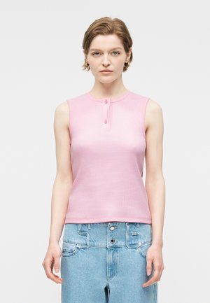 Νεαρή γυναίκα με κοντά μαλλιά που φοράει χρυσούς κρίκους στα αυτιά, ροζ αμάνικη μπλούζα με κουμπιά και ανοιχτόχρωμο τζιν ψηλόμεσο.