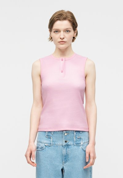 Jeune femme aux cheveux courts portant des boucles d'oreilles créoles dorées, un haut rose sans manches avec des boutons, et un jean bleu clair taille haute.