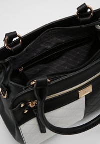 Valkoinen ja musta tikattu nahkalaukku, jossa on kultaiset yksityiskohdat, vetoketjutaskut ja musta kangasvuori, jossa on hienovarainen kuvio.