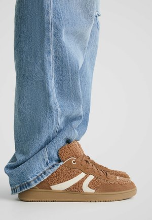 Persona que lleva vaqueros azul claro y zapatillas de forro polar marrón texturizadas con rayas beige, de pie sobre un fondo liso y claro.