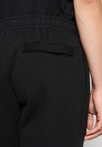Close-up of a person wearing black pants with an elastic waistband and a single slit back pocket, right hand relaxed beside the hip.