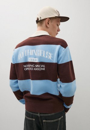 Ung mand iført en blå og brun stribet sweater med teksten "ANYTHING ELSE CLUB" og "NOTHING SPECIAL OPEN TO ANYONE" på ryggen.