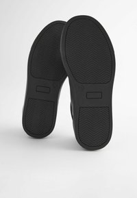 Semelles de chaussures en caoutchouc noir avec un motif en chevrons et des bords plats ; comprend un espace rectangulaire pour étiquetage près du talon.