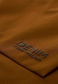 Smeđa teksturirana tkanina s suptilnim uzorkom rešetke. Crni logo "DENIM TOM TAILOR" istaknuto otisnut.